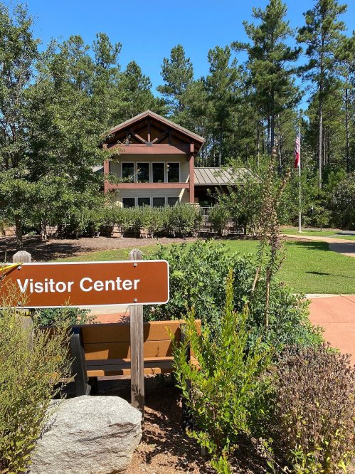 A visitor center surrounded by trees and greenery, with a sign in front and a clear blue sky above.