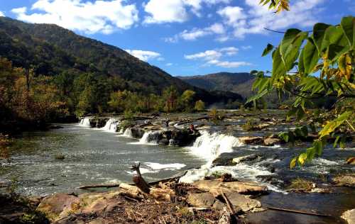 A serene river flows over rocks, surrounded by lush greenery and mountains under a blue sky with fluffy clouds.