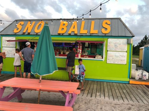 Colorful "Sno Balls" stand with people ordering, picnic tables in front, and string lights above on a beach boardwalk.