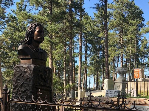 A bust of Calamity Jane stands in a cemetery surrounded by trees, with gravestones visible in the background.