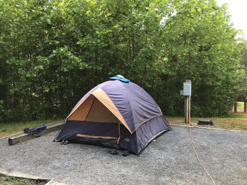 A camping tent set up on a gravel site, surrounded by green trees and a power outlet nearby.