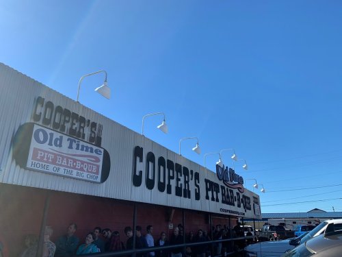 A busy barbecue restaurant with a long line of people outside under a clear blue sky.