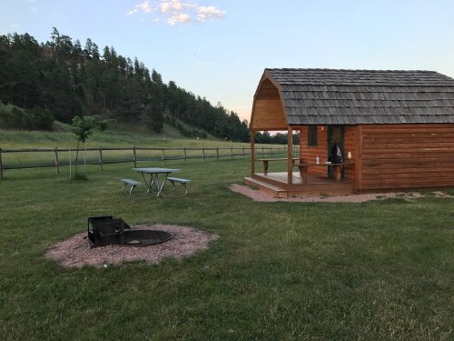 A wooden cabin with a porch sits in a grassy area, near a fire pit and picnic table, surrounded by trees.