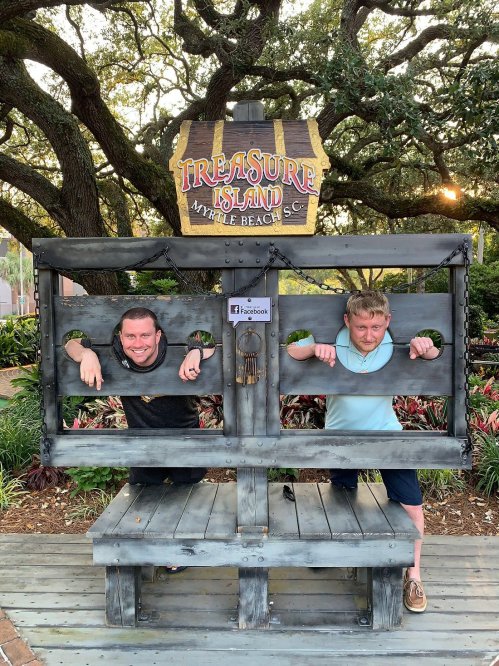 Two men pose in a wooden stocks display at Treasure Island in Myrtle Beach, surrounded by colorful plants and trees.