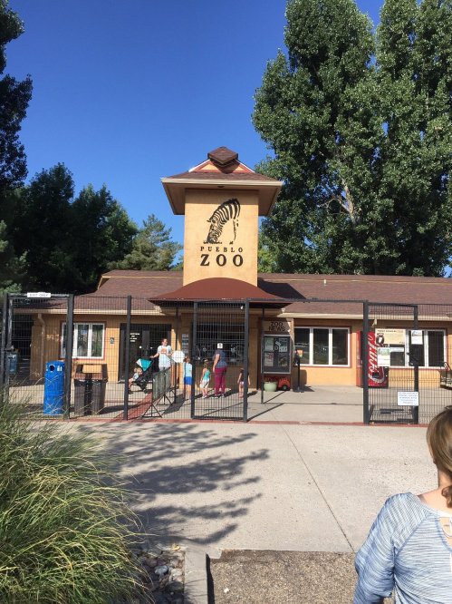 Entrance to Pueblo Zoo, featuring a large sign and visitors at the gate, surrounded by trees and blue sky.