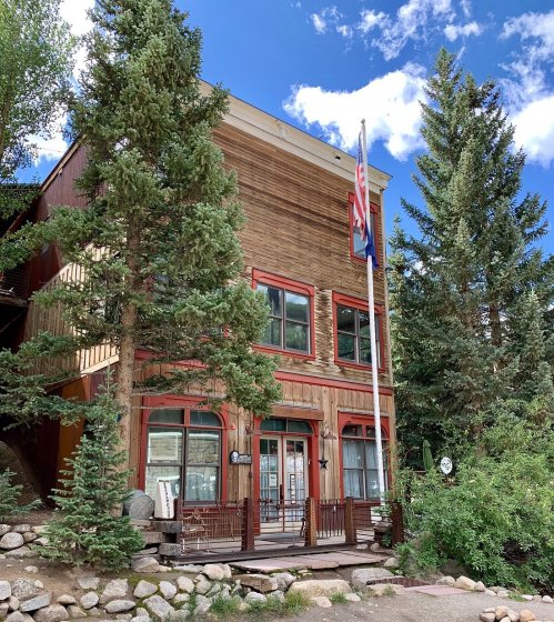 A wooden building surrounded by trees, featuring a flagpole with flags and a porch with seating. Blue sky above.