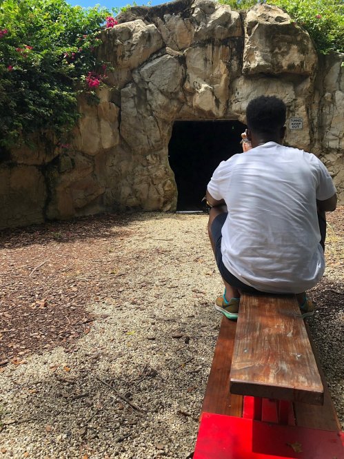 A person sits on a bench facing a dark cave entrance surrounded by greenery and flowers.