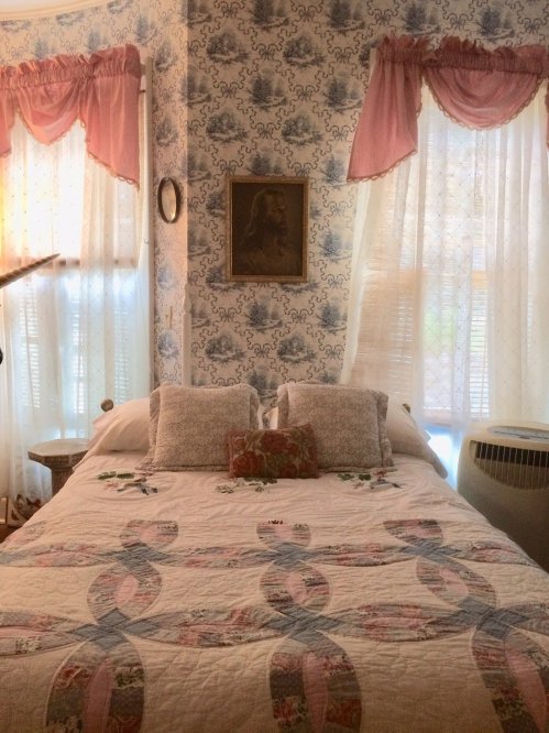 Cozy bedroom with a quilted bed, floral wallpaper, and soft curtains letting in natural light.