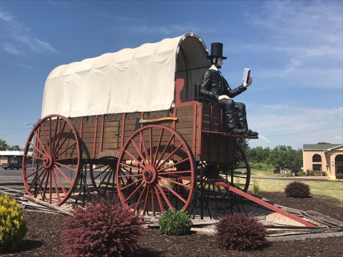 A large statue of a man in a top hat reading a book, seated on a covered wagon, surrounded by shrubs and blue sky.