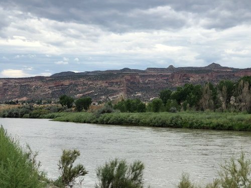 A river flows beside lush greenery, with rocky hills and cloudy skies in the background.