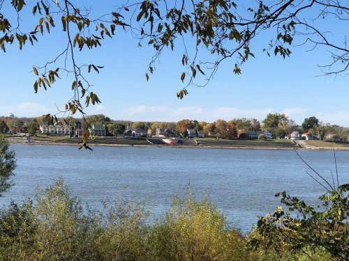 A serene river view with houses along the shore, framed by trees and a clear blue sky.