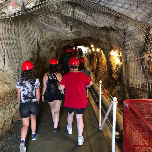 Visitors wearing hard hats walk through a dimly lit cave tunnel with rocky walls and safety railings.