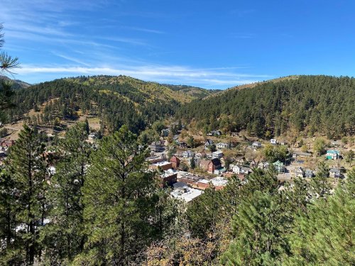 A scenic view of a small town nestled in green hills under a clear blue sky, surrounded by trees and mountains.