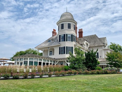 A large, elegant white house with a tower, surrounded by greenery and colorful flowers under a cloudy sky.