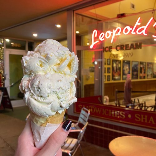 A hand holds a large ice cream cone outside Leopold's Ice Cream shop, with a neon sign in the background.