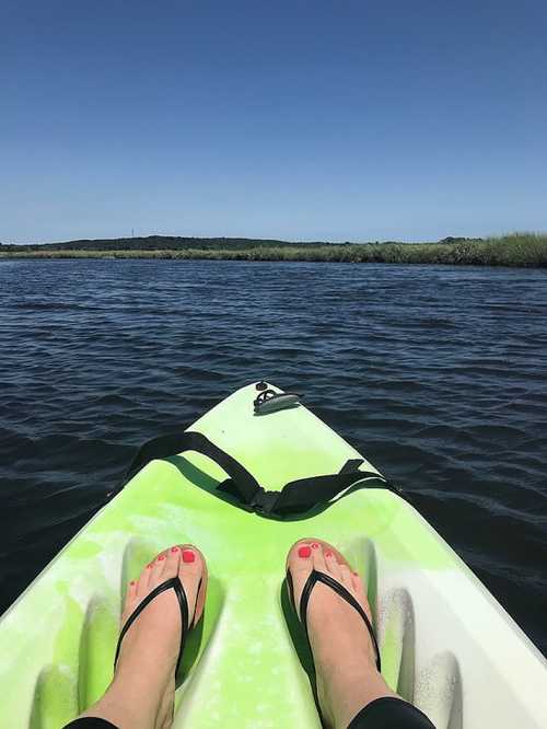 A person's feet in flip-flops resting on a kayak, with calm water and a clear blue sky in the background.