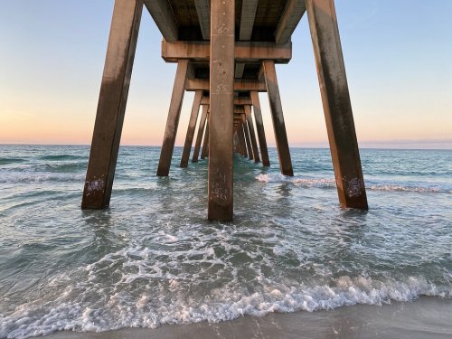 View from beneath a pier, with wooden supports rising above gentle waves and a pastel sky at sunset.