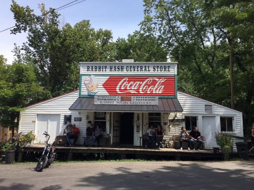 A rustic general store with a Coca-Cola sign, people sitting on the porch, surrounded by trees in Rabbit Hash, Kentucky.