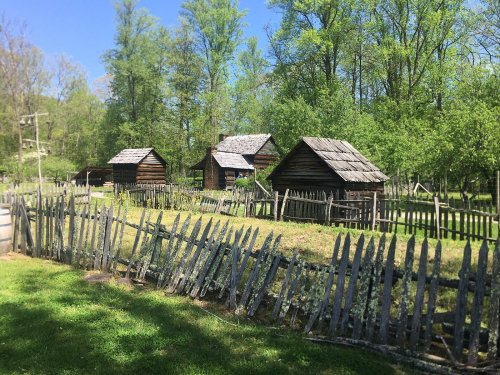 A rustic scene featuring three wooden cabins surrounded by a picket fence and lush green trees under a clear blue sky.