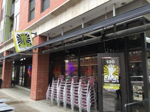 Exterior of a restaurant named "Zombica" with stacked chairs outside and colorful neon lights visible through the windows.