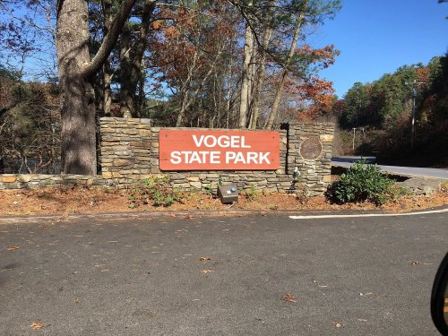 Sign for Vogel State Park, surrounded by trees and a clear blue sky, with a stone base and a paved road nearby.