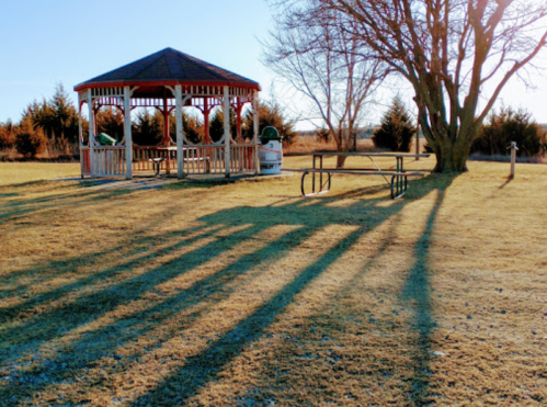 A gazebo with a picnic table casts long shadows on a grassy field under a clear blue sky.