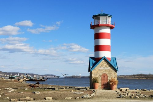 A red and white striped lighthouse stands by a river, surrounded by rocks and sculptures, under a blue sky with clouds.