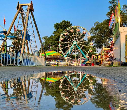 A carnival scene with a ferris wheel and rides reflected in a puddle on the ground under a clear blue sky.