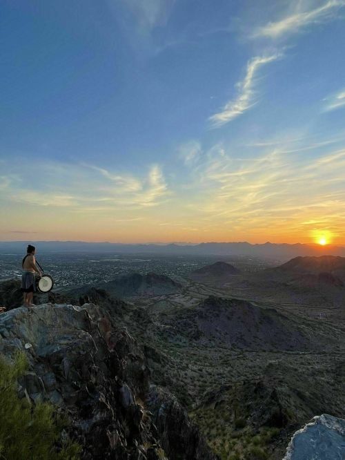 A person stands on a rocky ledge, playing a drum against a backdrop of a sunset over a vast mountainous landscape.