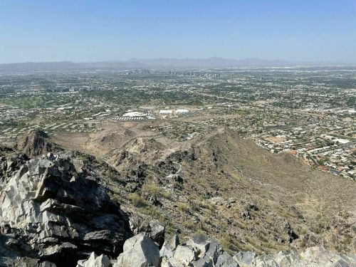 A panoramic view from a mountain peak overlooking a sprawling city and surrounding landscape under a clear blue sky.