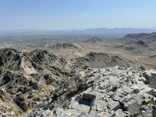 A panoramic view of rocky mountains and a valley under a clear blue sky, with distant hills on the horizon.