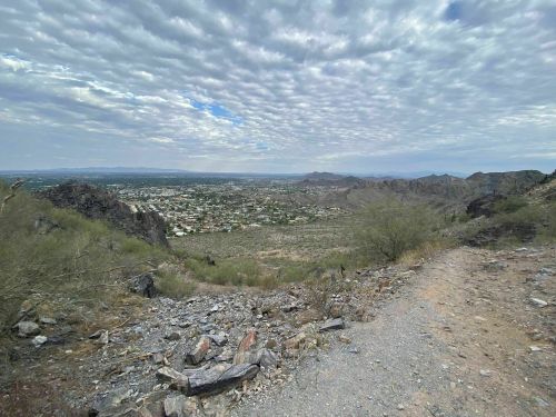 A rocky trail leads down a mountain, overlooking a valley and town under a cloudy sky.