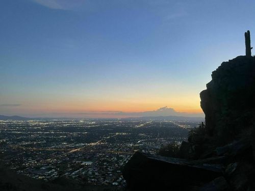 A scenic view of a city at dusk, with lights twinkling below and a rocky silhouette in the foreground.