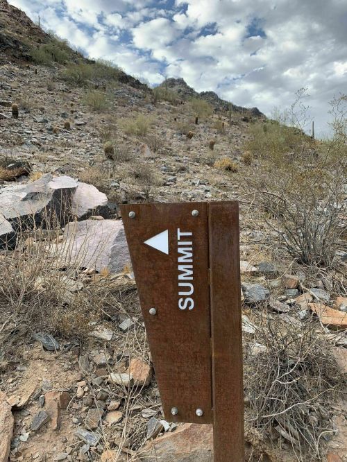 A weathered sign pointing to the summit, set against a rocky, arid landscape with scattered vegetation and clouds.
