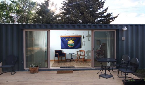 A cozy outdoor space with a Montana flag, chairs, and a table, framed by a shipping container and trees.
