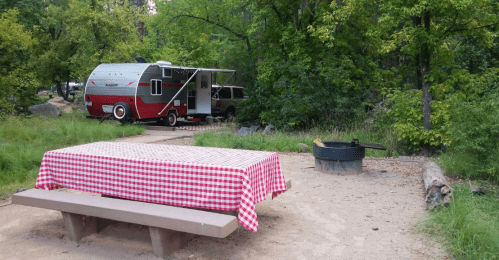 A vintage camper parked in a wooded area, with a picnic table covered in a red checkered tablecloth and a fire pit nearby.