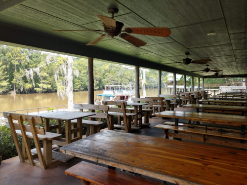 Outdoor dining area with wooden tables and fans, overlooking a river surrounded by trees.