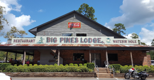 Exterior of Big Pines Lodge restaurant with a sign and steps, surrounded by greenery under a blue sky.