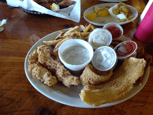 A plate of fried chicken tenders, fish fillet, French fries, and dipping sauces on a wooden table.