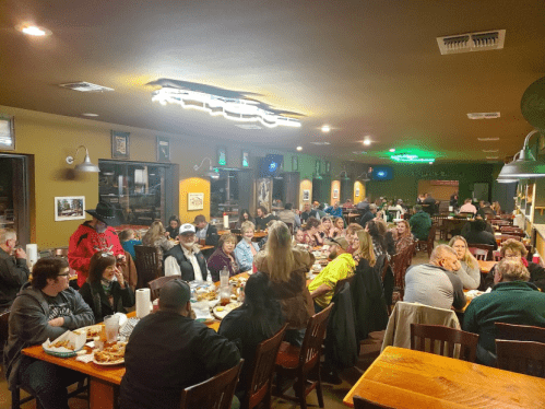 A crowded restaurant with people dining at long tables, enjoying food and drinks in a lively atmosphere.