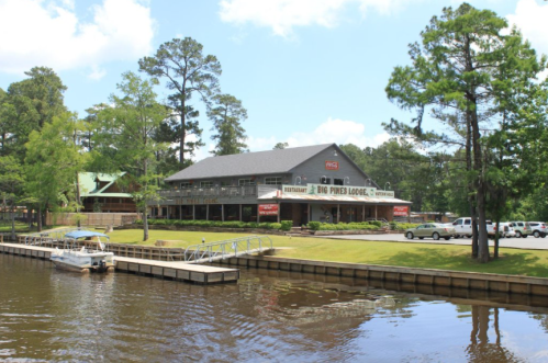 A rustic lodge by the water, surrounded by trees, with a dock and parking area in front. Bright blue sky above.