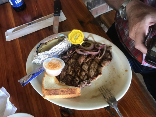 A plate with a grilled steak, baked potato, grilled onions, a slice of bread, and a dipping sauce.