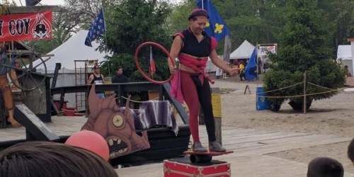 A performer balances on a box while juggling a hoop at an outdoor festival, with colorful flags and tents in the background.