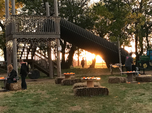 A playground with a slide, hay bales, and pumpkins at sunset, with people walking in the background.