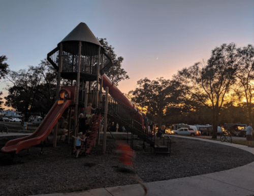 A playground with a large slide at sunset, children playing, and campers in the background.