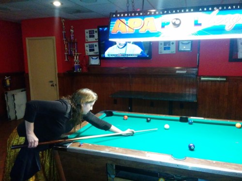 A woman in a black top and patterned skirt leans over a pool table, preparing to take a shot.