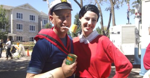 A man dressed as Popeye and a woman as Olive Oyl pose together, smiling, in a sunny outdoor setting.