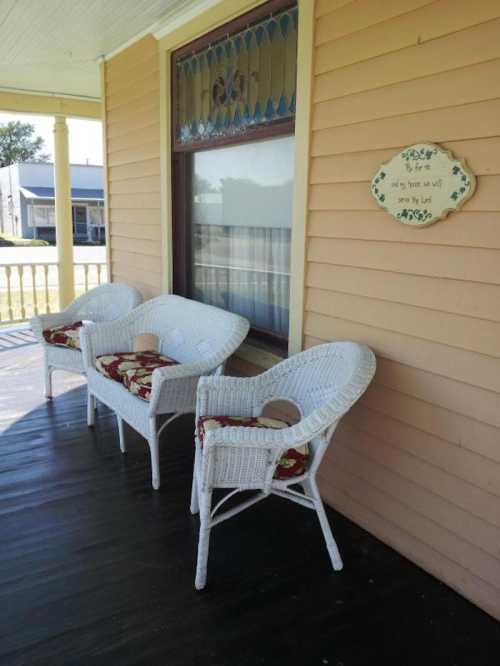 A porch with three white wicker chairs and decorative cushions, featuring a sign on the wall.