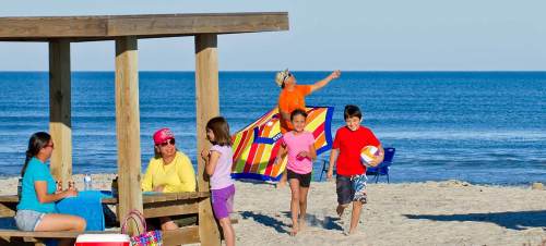 A sunny beach scene with children playing, a woman with a beach towel, and adults relaxing under a wooden shelter.