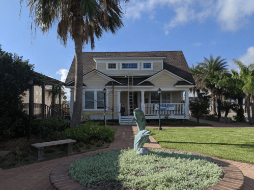 A charming two-story house with a porch, surrounded by palm trees and a landscaped garden featuring a sculpture.